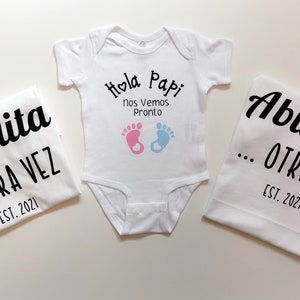May include: Three white t-shirts with black text. The baby onesie says "Hola Papi Nos Vemos Pronto" with pink and blue footprints. The other two shirts say "Abuelita...Otra Vez Est. 2021" and "Abuelito...Otra Vez Est. 2021".