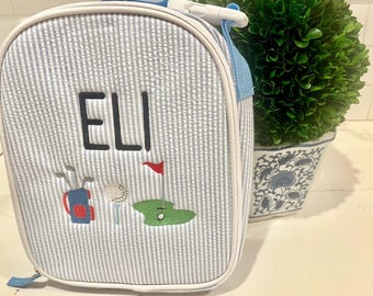 May include: A blue and white striped lunch bag with the name "ELI" embroidered on the front. The bag features a golf-themed design with a golf bag, golf balls, and a golf flag.