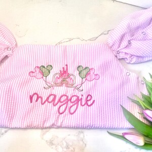 Puede incluir: Romper de bebé a rayas rosas y blancas con globos de Mickey Mouse bordados y un diseño de castillo. El nombre "Maggie" está bordado en rosa. El romper tiene mangas cortas y cierres a presión.