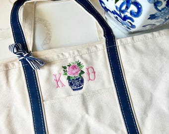 May include: A white canvas tote bag with navy blue trim and a pocket. The bag has a pink embroidered floral design with the initials "K.O.D."