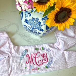 Puede incluir: Un sombrero de sol de seersucker rosa claro con iniciales bordadas y motivos marinos. El sombrero presenta un monograma en escritura rosa, rodeado de medusas y peces bordados. Un jarrón con girasoles y otras flores está en el fondo.