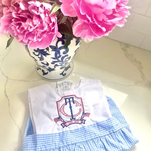 Könnte beinhalten: Ein weiß-blaues Gingham-Kleid mit einem weißen Oberteil, das mit einem rot-weiß-blauen gestickten Monogramm verziert ist. Das Monogramm ist ein Schild mit den Buchstaben "TC" darin. Das Kleid hat einen Rüschen-Saum.