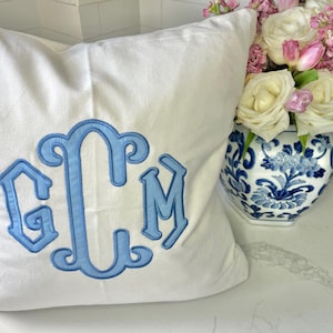 May include: A white square pillow with a light blue monogram of the letters GCM. The pillow is next to a blue and white floral vase filled with white and pink flowers.