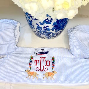 Puede incluir: Conjunto de gorro y babero para bebé a rayas azules y blancas. El babero presenta un barco, boyas y dos perros golden retriever. El conjunto está bordado con las iniciales "TJP" en rojo. El gorro tiene un borde con volantes.