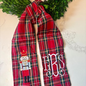 Puede incluir: Bufanda a cuadros rojos con diseño de cascanueces y monograma blanco. La bufanda presenta un patrón de tartán clásico en rojo, verde y marrón. Un cascanueces está bordado en un extremo y un monograma en el otro.