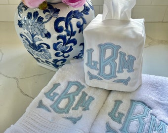 May include: Two white hand towels with blue embroidered initials "LBM" and a white tissue box cover with the same blue embroidered initials. The tissue box cover is sitting on top of the towels.