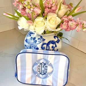 May include: A blue and white striped cosmetic bag with a monogrammed initial "GG" in a blue floral wreath. The bag is sitting on a white countertop in front of a vase of white and pink flowers.