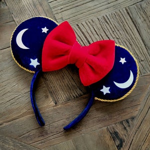 May include: Blue velvet Minnie Mouse ears with a red velvet bow and gold trim. The ears feature white stars and crescent moons.