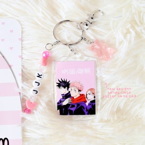 May include: A clear acrylic keychain featuring a Jujutsu Kaisen anime design with three characters. The keychain includes a silver clasp, a pink gummy bear charm, and a beaded JJK name tag. The background is a fluffy white surface.