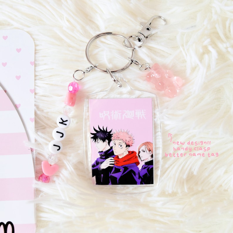 May include: A clear acrylic keychain featuring a Jujutsu Kaisen anime design with three characters. The keychain includes a silver clasp, a pink gummy bear charm, and a beaded JJK name tag. The background is a fluffy white surface.