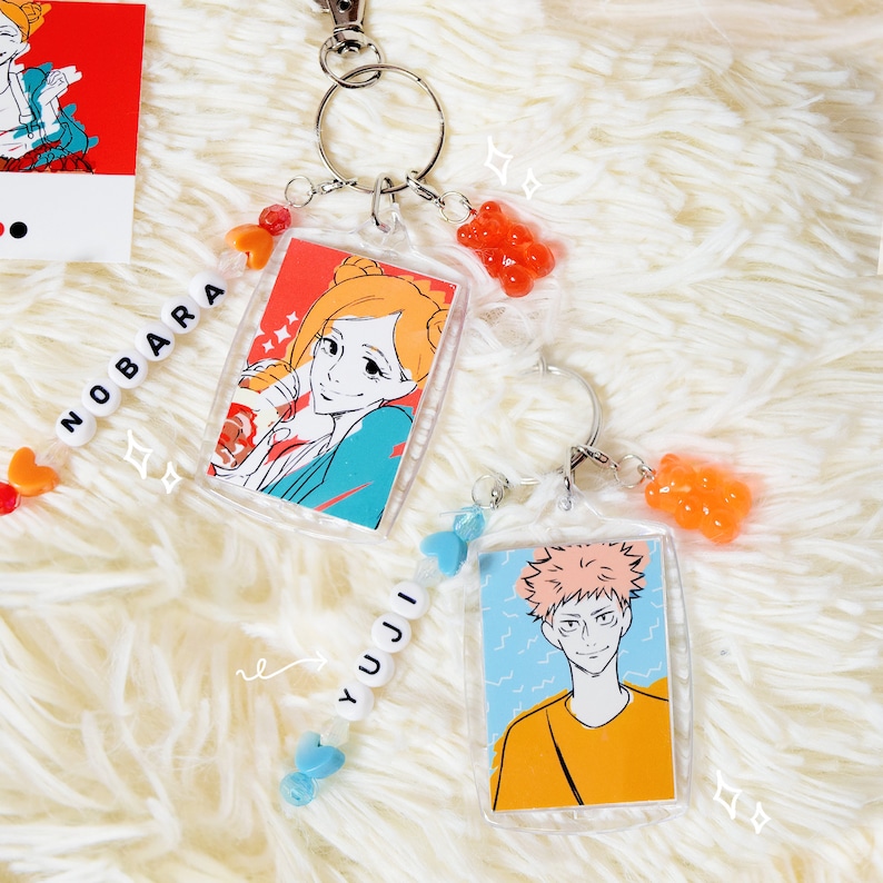 May include: Clear acrylic keychains featuring anime characters. One keychain has a character with orange hair and the name "NOBARA" spelled out in white beads. The other keychain has a character with pink hair and the name "YUJI" in white beads.
