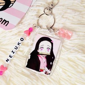 May include: A clear acrylic keychain featuring a Nezuko Kamado anime character illustration. The keychain includes a silver clasp, a pink gummy bear charm, and a beaded lanyard spelling "NEZUKO" with pink heart beads.