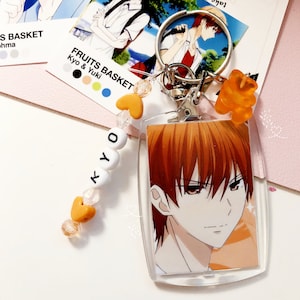 May include: Clear acrylic keychain featuring an anime character with orange hair and red eyes. The keychain has a silver clasp, orange heart-shaped beads, and the name "KYO" spelled out in white letter beads. A small orange gummy bear charm is also attached.