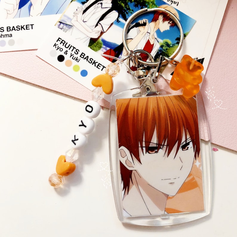 May include: Clear acrylic keychain featuring an anime character with orange hair and red eyes. The keychain has a silver clasp, orange heart-shaped beads, and the name "KYO" spelled out in white letter beads. A small orange gummy bear charm is also attached.
