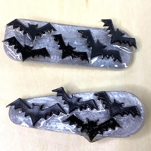 Puede incluir: Dos decoraciones ovaladas con base gris y diseños de murciélagos negros. Cada artículo presenta varias siluetas de murciélagos negros, creando un tema de Halloween o gótico. Los murciélagos están dispuestos sobre la superficie.