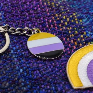 May include: A keychain and a patch featuring the non-binary pride flag. The keychain has a circular charm with the flag's colors: yellow, white, purple, and black. The patch is a rainbow design with the same colors. Both items are on a textured purple background.