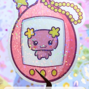 Może przedstawiać: Holograficzna naklejka w kolorze r&oacute;żowym i ż&oacute;łtym przedstawiająca cyfrowego zwierzaka w stylu Tamagotchi z r&oacute;żowym zwierzątkiem w środku. Naklejka ma gwiazdę na g&oacute;rze i ż&oacute;łtą pętlę łańcucha.