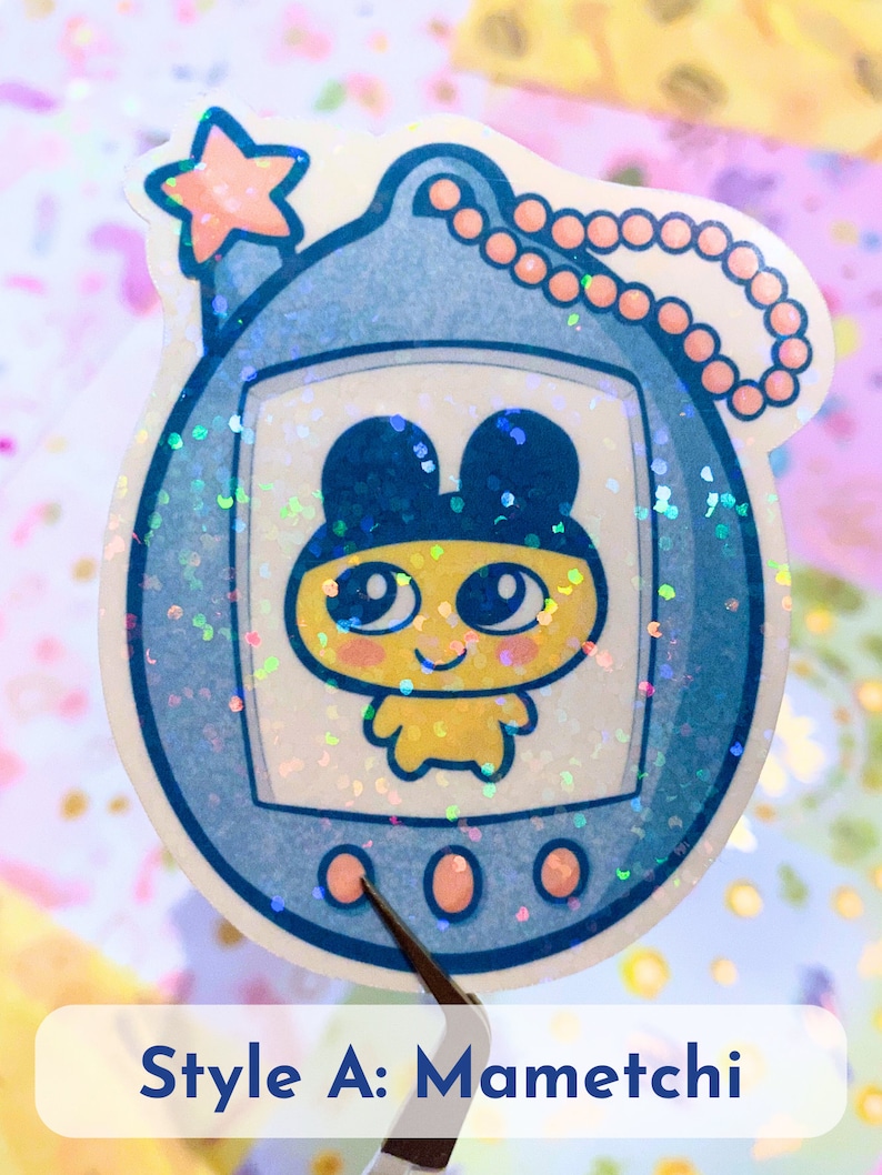 Może przedstawiać: Brelok w stylu Tamagotchi w kolorze niebieskim i białym z r&oacute;żową gwiazdką na g&oacute;rze i r&oacute;żowym łańcuszkiem. Na ekranie widoczna jest ż&oacute;łta postać z czarnymi uszami i dużymi oczami. Pod brelokiem widoczny jest tekst "Style A: Mametchi".