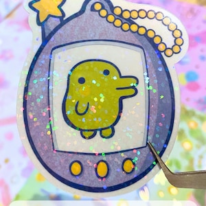 Może przedstawiać: Naklejka z brokatem w kolorze fioletowym i ż&oacute;łtym przedstawiająca Tamagotchi z ż&oacute;łtym pisklęciem na ekranie. Naklejka ma złoty łańcuszek przymocowany do g&oacute;rnej części.