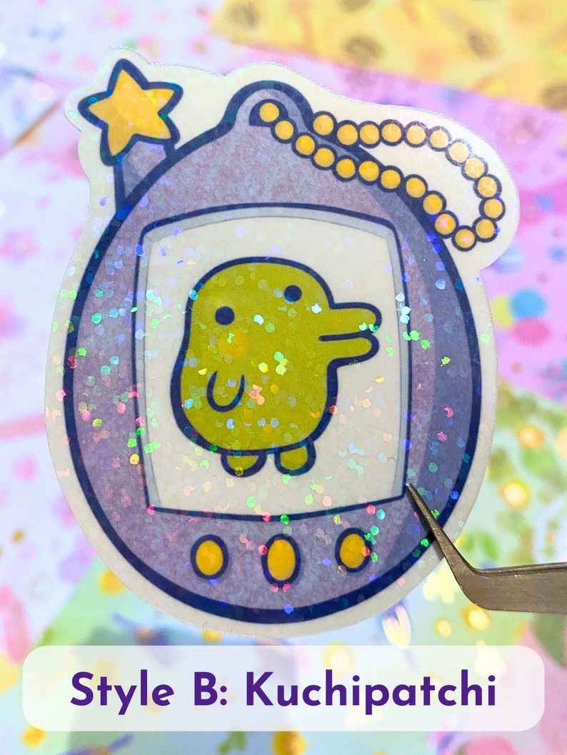 Może przedstawiać: Naklejka z brokatem w kolorze fioletowym i ż&oacute;łtym przedstawiająca Tamagotchi z ż&oacute;łtym pisklęciem na ekranie. Naklejka ma złoty łańcuszek przymocowany do g&oacute;rnej części.