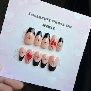 May include: A set of pink press-on nails with black tips and gold glitter accents. Each nail features a red heart with a gold cross and a gold star design. The nails are displayed on a white card with the text "Colleen's Press On Nails".