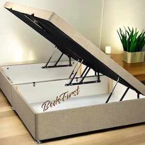 May include: A beige upholstered bed frame with a lift-up storage compartment. The frame is open, revealing the storage space and the black gas lift mechanism. The text "BedsFirst" is visible on the frame.