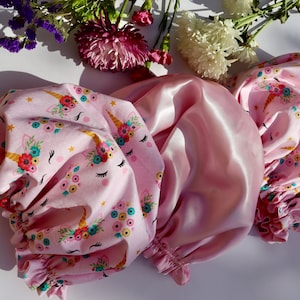 May include: Two pink satin hair bonnets with a unicorn and floral print. One bonnet is made of plain satin and the other is made of satin with a printed design.