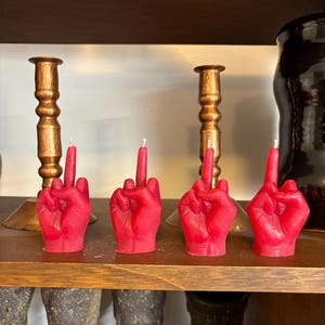 May include: Four red candle sculptures shaped like a middle finger. The candles are on a wooden shelf with copper candle holders.