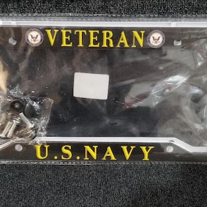 May include: A black license plate frame with the words "VETERAN" and "U.S. NAVY" in yellow. The frame has a silver border and includes hardware. The packaging has a small American flag design.