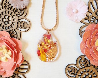 May include: A clear teardrop-shaped pendant necklace with gold flakes and dried flowers encased in resin. The pendant hangs from a delicate gold chain. The background features paper flowers and wooden cutouts in shades of pink and peach.