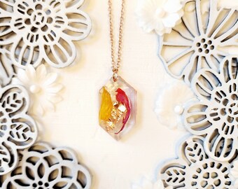 May include: A rose gold necklace with a clear, hexagonal pendant. The pendant contains yellow and red dried flowers and gold leaf. The background features white floral decorations and cut-out designs.