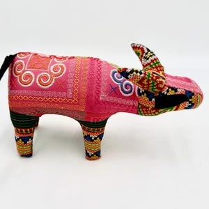 May include: A handcrafted, stuffed animal in the shape of a water buffalo. The plush toy is primarily pink with colorful embroidered patterns. It has a black tail with white fringe and multi-colored horns.