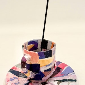 May include: A colorful, handmade teacup and saucer set with a patchwork design. The cup and saucer feature a mix of pink, purple, yellow, and blue hues. A black incense stick is placed inside the cup. The set is ideal for home decor.