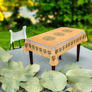May include: A rectangular mustard-colored tablecloth with a floral pattern and a decorative border. The tablecloth is draped over a dark brown wooden table. Large green leaves are scattered around the table.