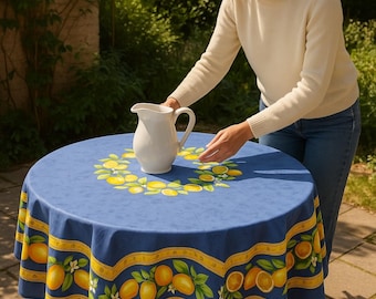 May include: A round blue tablecloth featuring a central wreath of yellow lemons and green leaves, with a wide yellow border decorated with a repeating pattern of lemons and foliage, topped with a plain white ceramic pitcher.