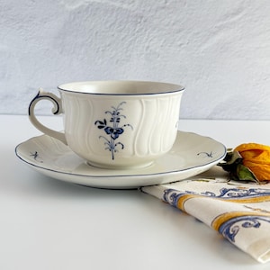 May include: A white teacup and saucer set with blue floral detailing. The cup has a curved handle and a blue trim. The saucer has a matching blue trim and floral accents. A yellow rose and patterned napkin are also present.