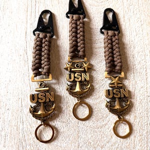 May include: Three keychains with brown braided cords and black clasps. Each keychain features a gold-colored anchor pendant with the letters "USN" and a circular key ring. One keychain has stars.