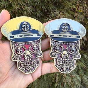 May include: Two decorative coins featuring a skull design with a naval officer's hat. The coins have a gold and silver finish, with the text "USN" and "CLASS 132 FY26" visible. The skull has a red and purple color scheme.