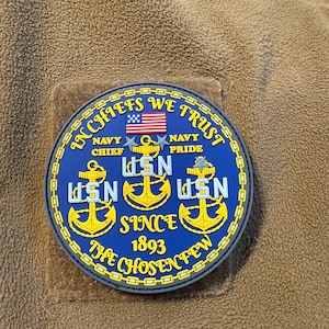May include: A round, blue patch with gold trim and text. The patch features the words "IN CHIEFS WE TRUST", "NAVY CHIEF PRIDE", and "USN" with anchors. An American flag is at the top, and the text "SINCE 1893 THE CHOSEN FEW" is at the bottom.