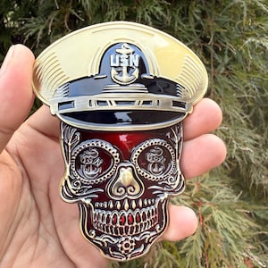 May include: A gold-colored metal skull-shaped coin with a red center and a gold-colored hat on top. The hat has the letters "USN" and an anchor emblem. The skull has intricate details and the coin is held in a hand.
