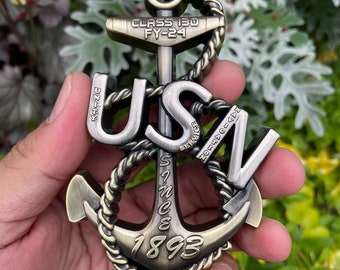 May include: A gold-toned metal anchor charm with the words "USN" and "Since 1893" inscribed on it. The anchor is wrapped with a rope design and has a small tag that reads "Class 130 FY-24".