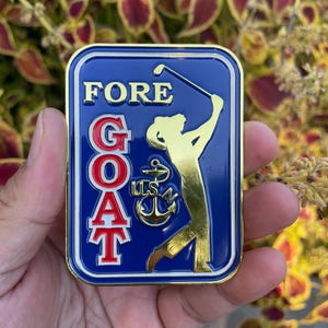 K&ouml;nnte beinhalten: Eine goldfarbene Metallstifte mit blauem Hintergrund und einer Golfer-Silhouette. Die Stifte sagt "FORE GOAT" in roten und wei&szlig;en Buchstaben mit einem goldenen Anker und "USN" in der Mitte.