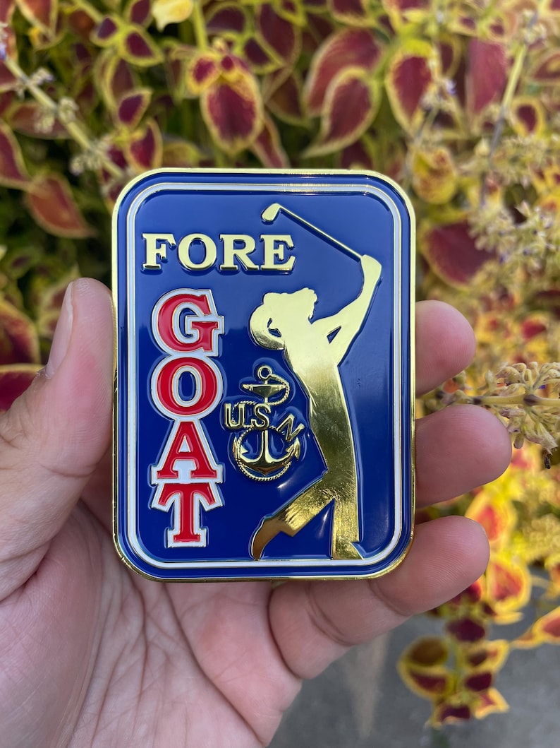 K&ouml;nnte beinhalten: Eine goldfarbene Metallstifte mit blauem Hintergrund und einer Golfer-Silhouette. Die Stifte sagt "FORE GOAT" in roten und wei&szlig;en Buchstaben mit einem goldenen Anker und "USN" in der Mitte.