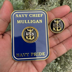 K&ouml;nnte beinhalten: Eine goldfarbene Metallplakette mit blauem Hintergrund und goldener Beschriftung. Die Plakette tr&auml;gt die Aufschrift "Navy Chief Mulligan Navy Pride". Au&szlig;erdem ist eine goldfarbene Metallnadel mit schwarzem Hintergrund und goldener Beschriftung zu sehen. Die Nadel tr&auml;gt die Aufschrift "USN".