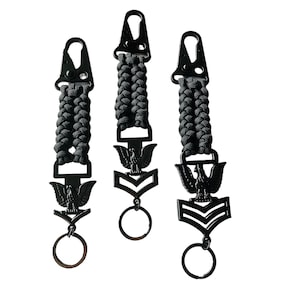 May include: Three black keychains with a braided cord design. Each keychain features a black metal clip, a decorative emblem with an eagle, and a ring. The keychains are designed with military insignia.