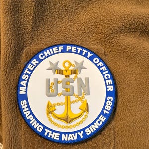 May include: A brown patch with the words "MASTER CHIEF PETTY OFFICER" in blue around the edge. The center features a gold anchor, the letters "USN", and the text "SHAPING THE NAVY SINCE 1893".