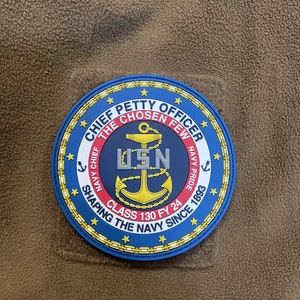 May include: A blue and gold embroidered patch with the words "Chief Petty Officer" and "The Chosen Few" around a gold anchor with the letters "USN" above it. The patch also includes the words "Navy Chief", "Navy Pride", "Class 130 FY 24", and "Shaping the Navy Since 1893".