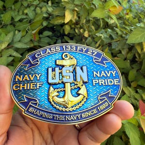 May include: An oval-shaped metal belt buckle with a blue diamond-plate background. It features a gold anchor, the letters USN, and text reading CLASS 133 FY 27, NAVY CHIEF, NAVY PRIDE, and SHAPING THE NAVY SINCE 1893, all framed by a gold-colored border.