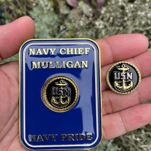 K&ouml;nnte beinhalten: Ein goldfarbenes Metallabzeichen mit blauem Hintergrund und wei&szlig;en Buchstaben. Das Abzeichen tr&auml;gt die Aufschrift "NAVY CHIEF MULLIGAN NAVY PRIDE". Ein goldfarbener Anker mit den Buchstaben "USN" befindet sich in der Mitte des Abzeichens. Eine kleinere goldfarbene Anstecknadel mit schwarzem Hintergrund und goldfarbenem Anker sowie den Buchstaben "USN" ist ebenfalls zu sehen.