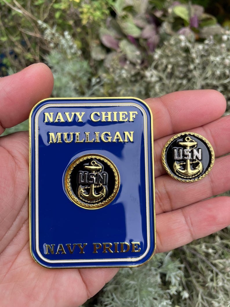 K&ouml;nnte beinhalten: Ein goldfarbenes Metallabzeichen mit blauem Hintergrund und wei&szlig;en Buchstaben. Das Abzeichen tr&auml;gt die Aufschrift "NAVY CHIEF MULLIGAN NAVY PRIDE". Ein goldfarbener Anker mit den Buchstaben "USN" befindet sich in der Mitte des Abzeichens. Eine kleinere goldfarbene Anstecknadel mit schwarzem Hintergrund und goldfarbenem Anker sowie den Buchstaben "USN" ist ebenfalls zu sehen.